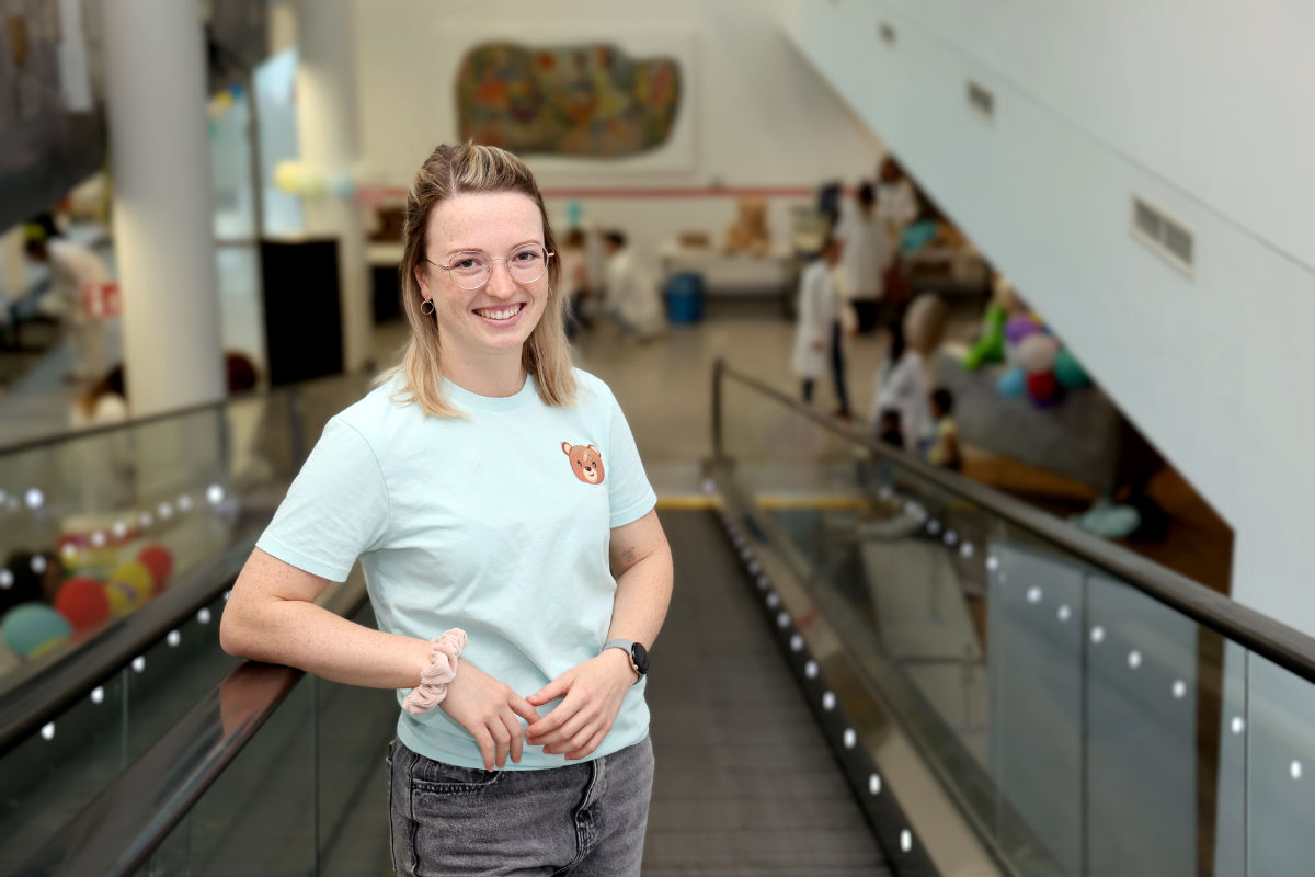 Medizinstudentin Claudia Hawlitzky im Teddybär-T-Shirt steht lächelnd im Klinikgebäude, im Hintergrund läuft das Teddybärkrankenhaus mit Kindern und HelferInnen.