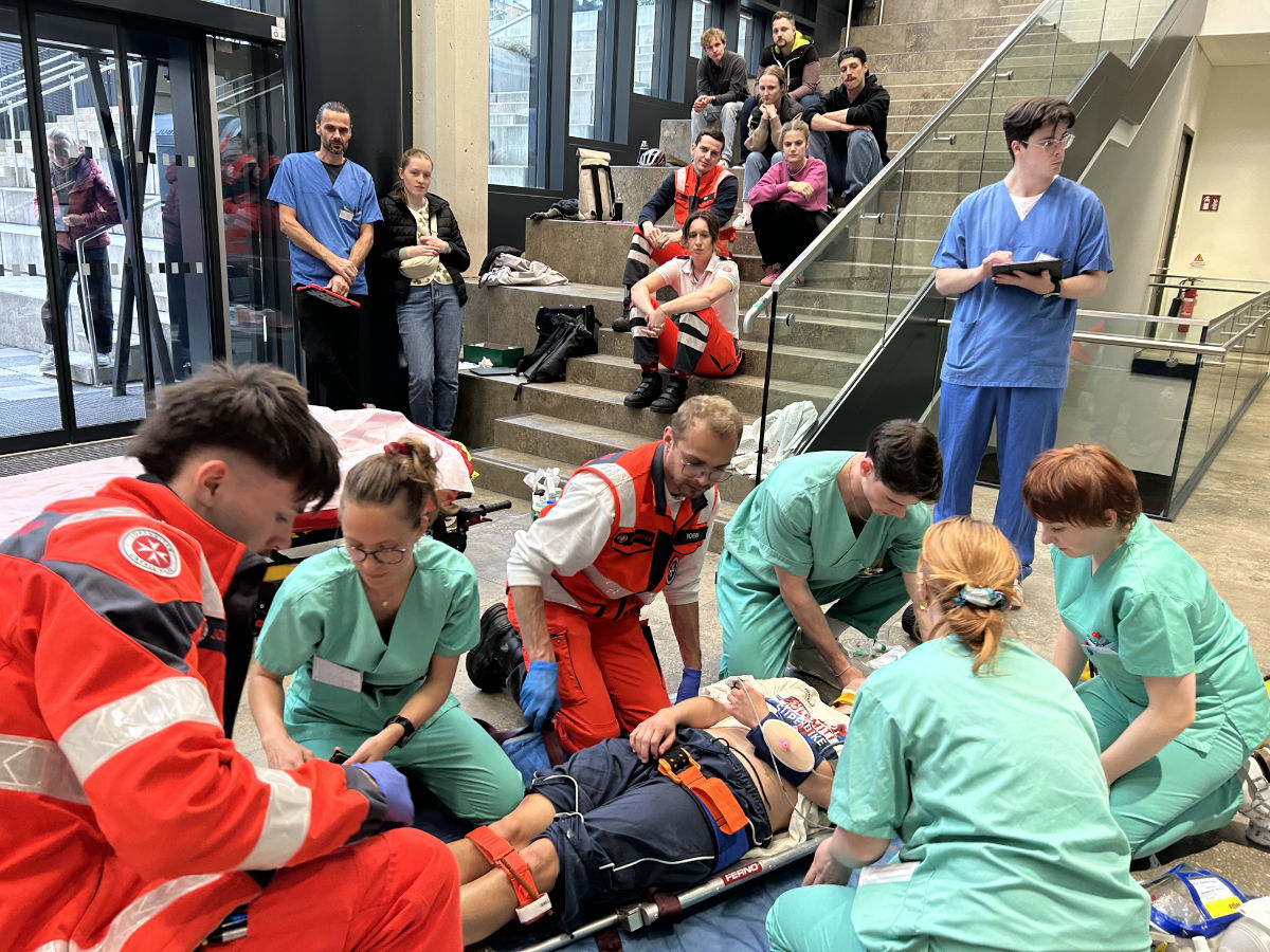 Vier Studierende und zwei Sanitäter kümmern sich um eine Patientin, die auf einer Trage liegt. Im Hintergrund wird die Übung von TutorInnen und ZuschauerInnen beobachtet.