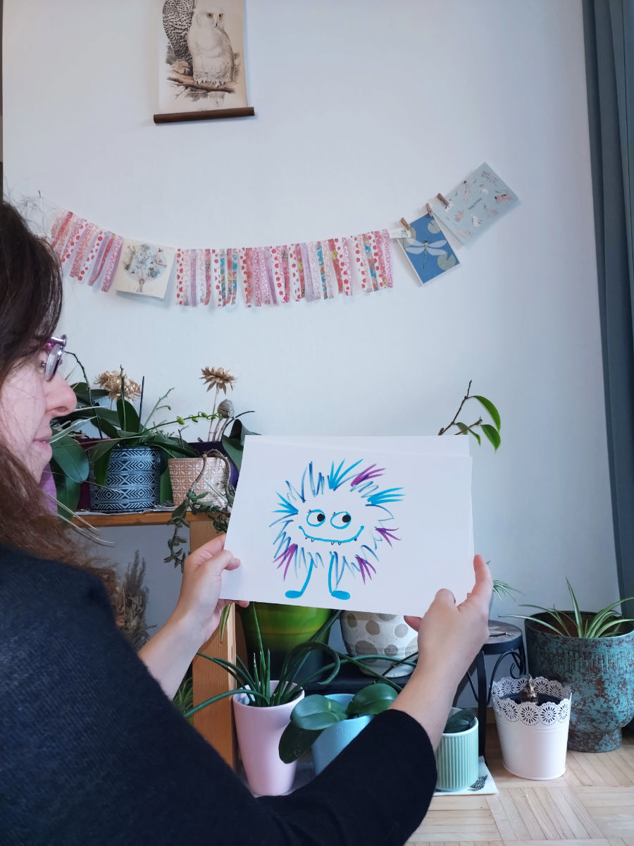 Birgit Sailer hält ein Blatt in der Hand, auf dem eines ihrer bunten, fröhlichen Monster zu sehen ist