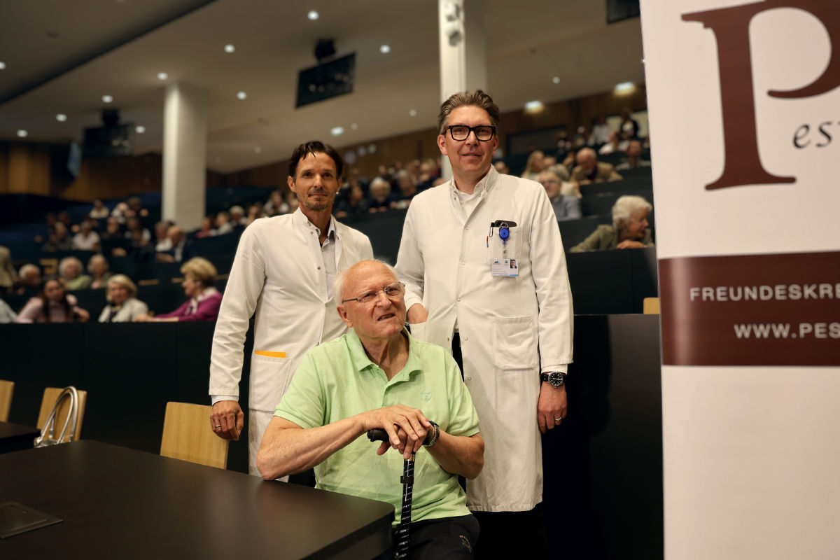 Raimund Margreiter im grünen T-Shirt sitzend, hinter stehen seine Schüler Stefan Schneeberger (l.) und Gerald Brandacher (r.), hinter ihnen die BesucherInnen im Hörsaal.