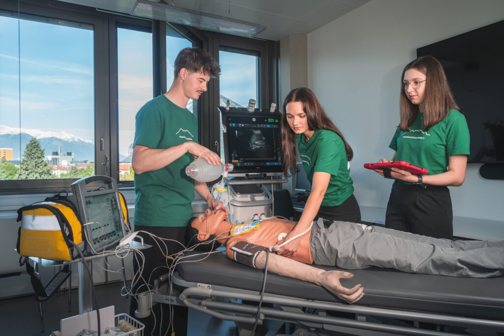 Drei Studierende bei einer Simulationsübung an einer Puppe: Diese liegt auf der Trage, ein Student beatmet sie, eine Studentin führt einen Ultraschal durch, eine Studentin steht mit einem Tablet dabei